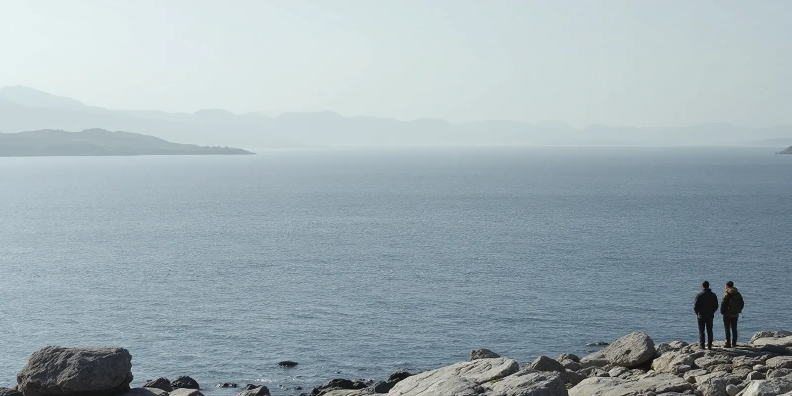 Estrecho gris azulado visto desde una orilla rocosa, con la costa de enfrente en el horizonte